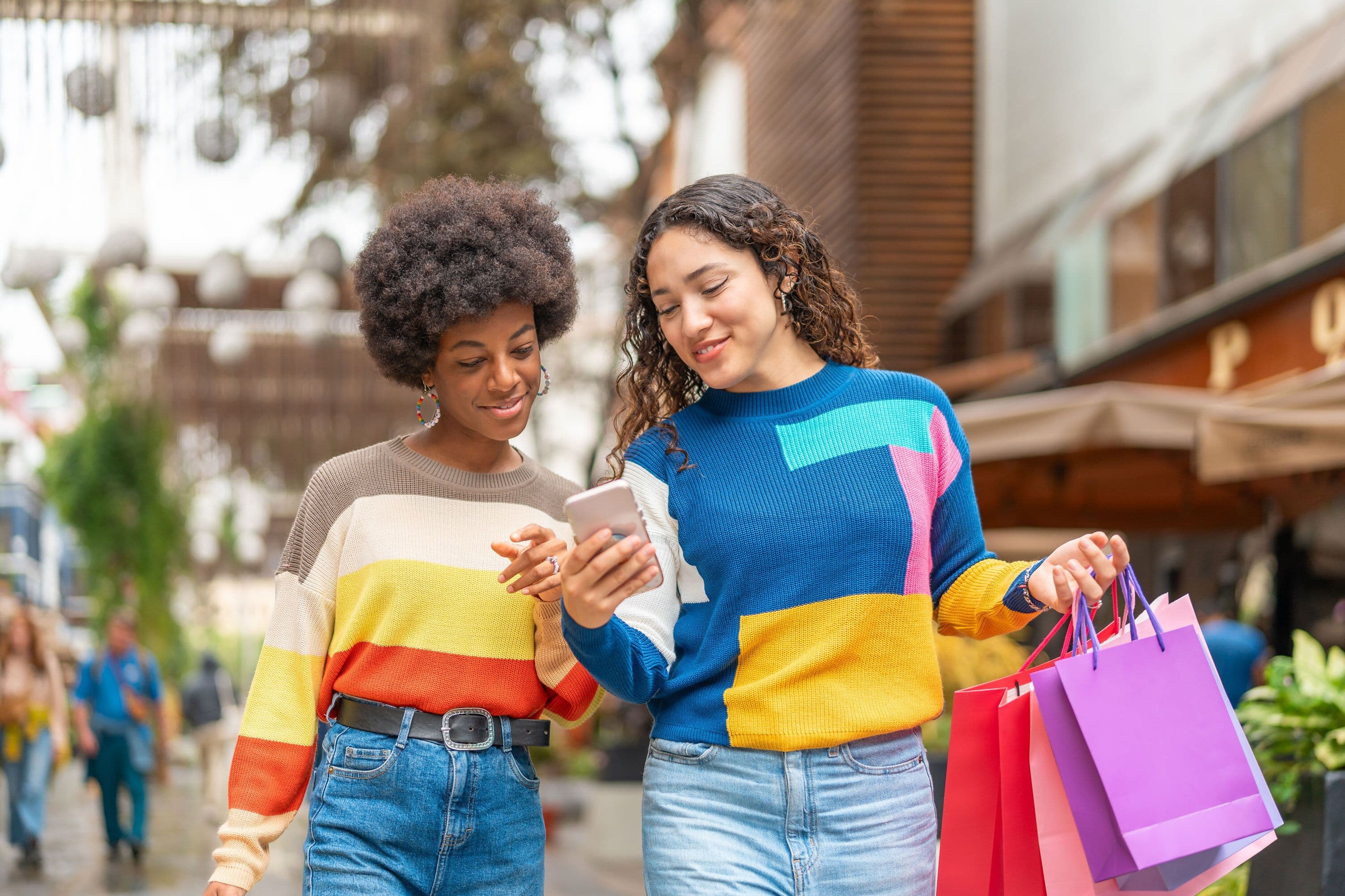 Two women in colorful sweaters walk with shopping bags, smiling and looking at a phone, in a lively outdoor setting.